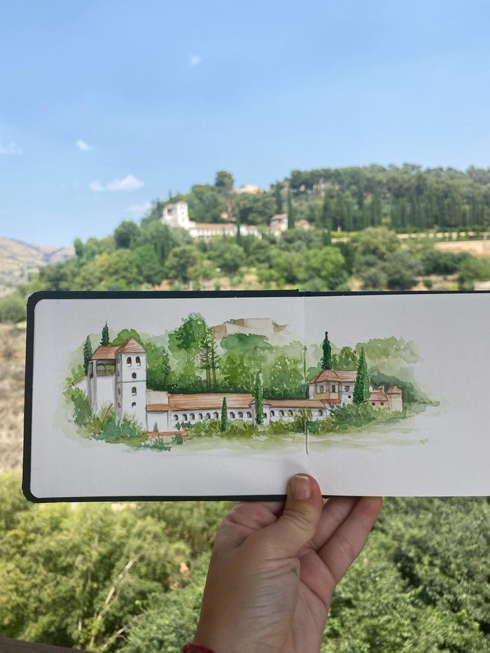 Sketchbook con Granada en acuarela, sostenido frente a la colina arbolada y sus edificios blancos